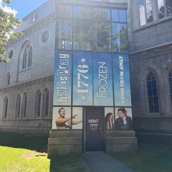 A stone building with large glass windows displaying posters for "Hairspray," "1776," "Frozen," and "Come From Away" musicals.