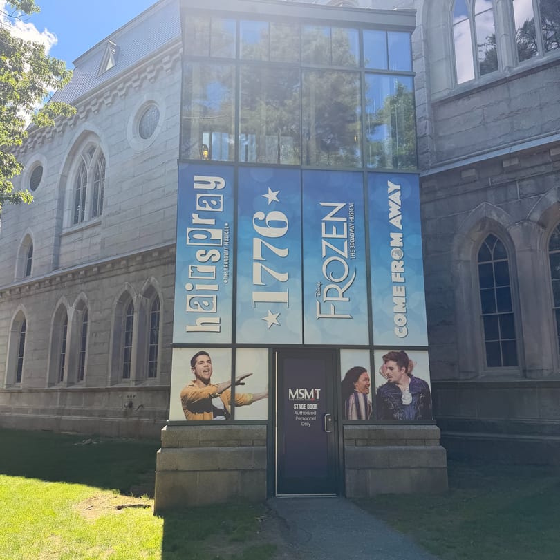 A stone building with large glass windows displaying posters for "Hairspray," "1776," "Frozen," and "Come From Away" musicals.