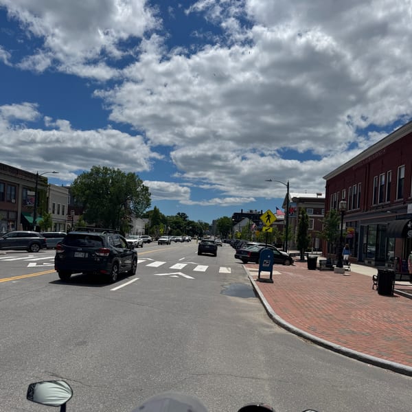 A bustling street scene with cars driving on a road lined with brick buildings and sidewalks under a cloudy sky.