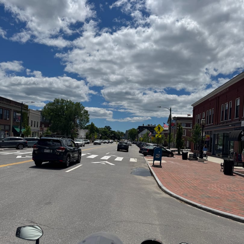 A bustling street scene with cars driving on a road lined with brick buildings and sidewalks under a cloudy sky.