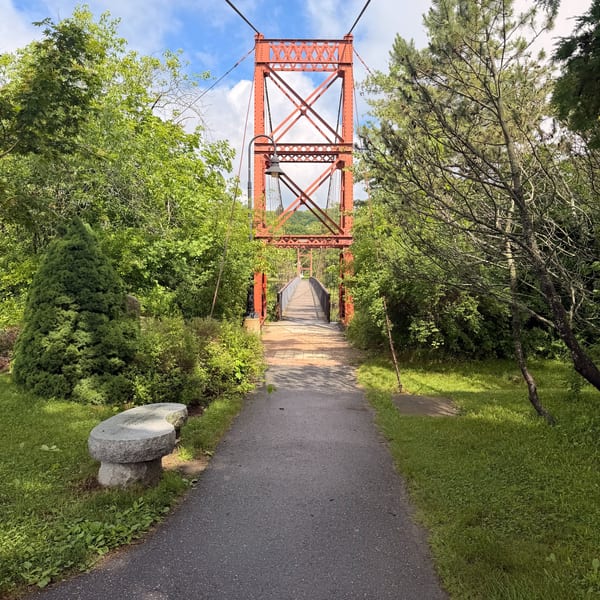 A red suspension bridge tower stands over a paved path surrounded by lush green trees and grass.
