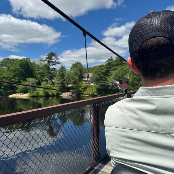 A person in a black hat and light shirt looks out from a bridge over a river.